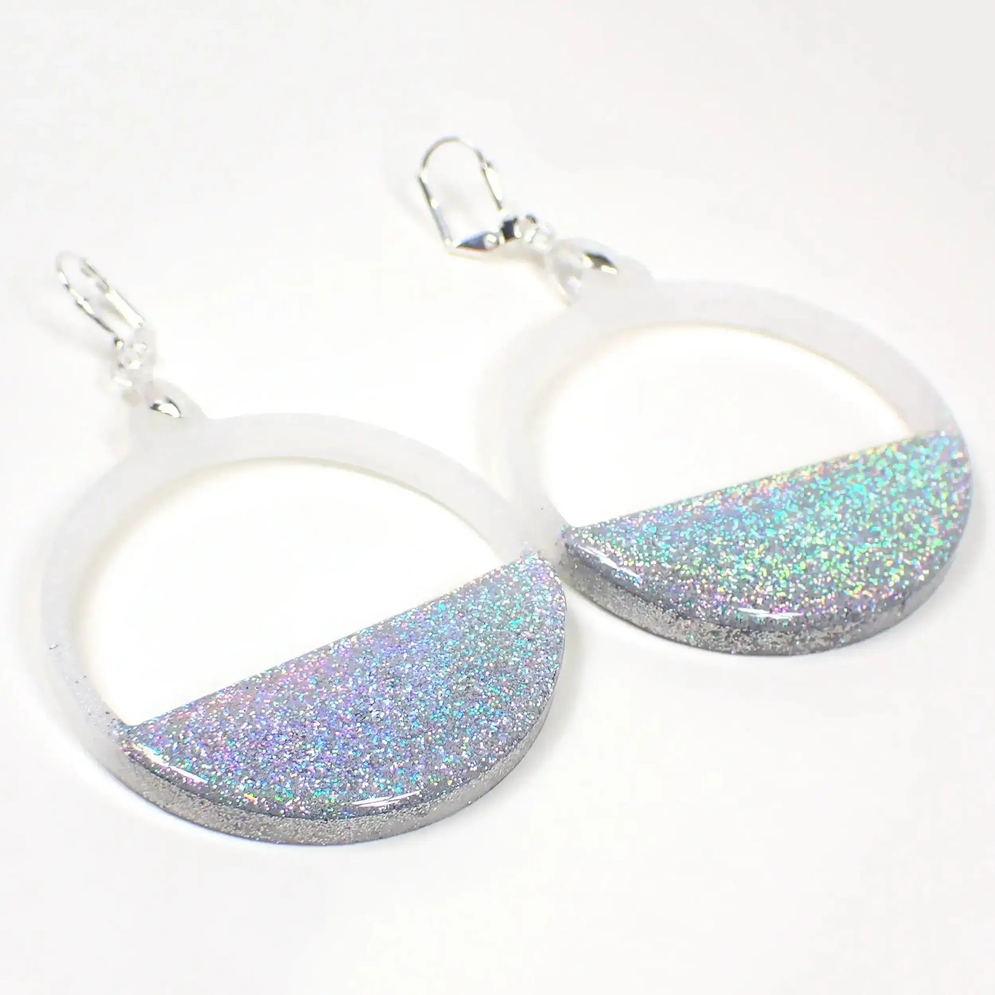 Angled front view of the handmade resin hoop earrings. The metal is silver tone plated in color. They are round hoop shaped with an open top and a semi circle bottom. The top part is pearly white in color. The bottom has silver holographic glitter embedded in it which has sparkle and flashes of different colors.