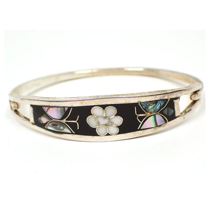 Front view of the retro vintage Taxco hinged bangle bracelet. The silver tone metal is slightly darkened from age. There is black enamel on the front with a white flower in the middle. Each side has a butterfly design made up of pieces of inlaid abalone shell.