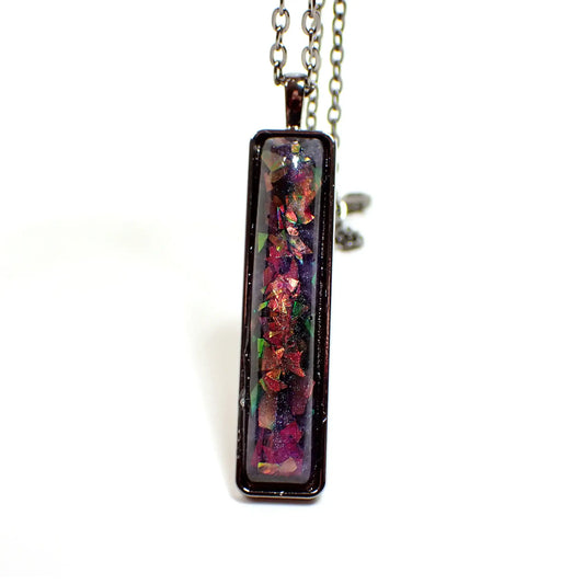 Enlarged front view of the handmade resin bar pendant necklace. The metal is gunmetal gray in color. The pendant is a long rectangle bar shape with rounded corners. The resin cab has marbled shades of pearly purple resin. There are chunky pieces of iridescent glitter with flashes of different colors as you move around in the light.