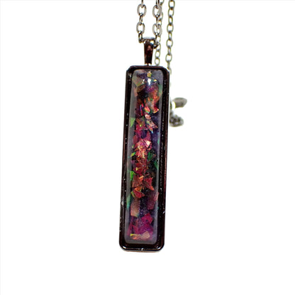 Enlarged front view of the handmade resin bar pendant necklace. The metal is gunmetal gray in color. The pendant is a long rectangle bar shape with rounded corners. The resin cab has marbled shades of pearly purple resin. There are chunky pieces of iridescent glitter with flashes of different colors as you move around in the light.