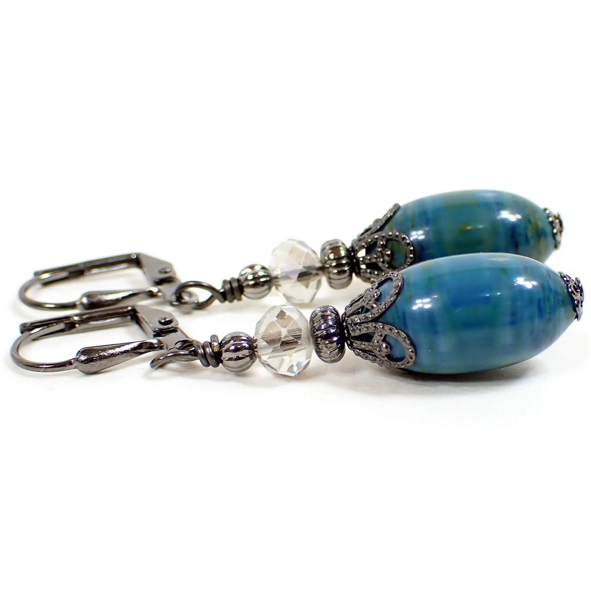 Side view of the handmade earrings with vintage lucite beads. The metal is gunmetal gray in color. There are faceted glass crystal beads at the top in a very light gray color. The bottom lucite beads are oval shaped and have marbled areas with different shades of blue and green. Each beads has a different pattern for a unique appearance.