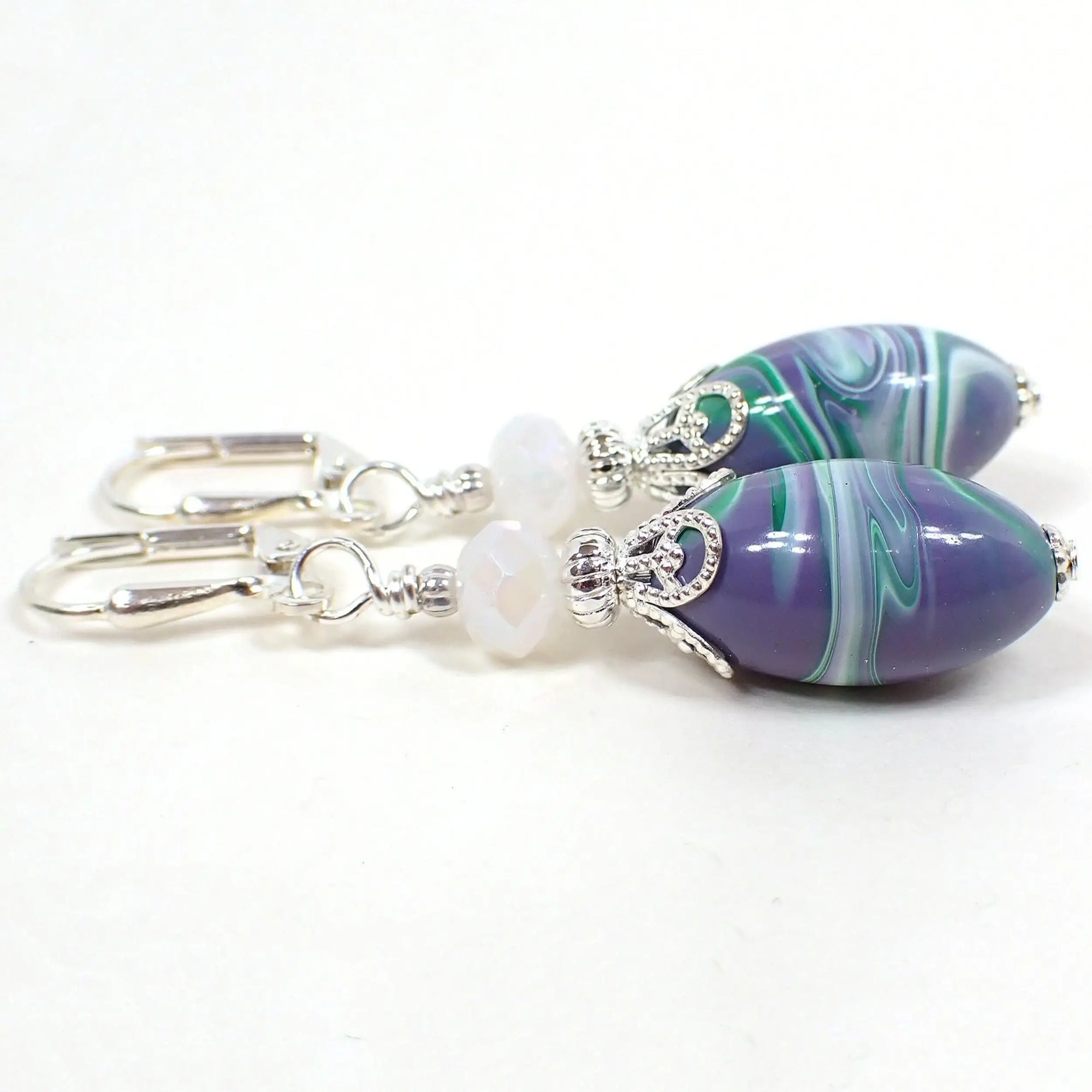 Side view of the handmade earrings with vintage lucite beads. The metal is silver plated in color. There are pearly opaque white faceted glass beads at the top. The bottom lucite beads are oval shaped and have marbled swirls of purple, green, and white. Each bottom bead has a different pattern than the other for a unique appearance.