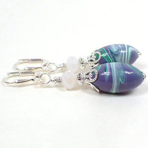Side view of the handmade earrings with vintage lucite beads. The metal is silver plated in color. There are pearly opaque white faceted glass beads at the top. The bottom lucite beads are oval shaped and have marbled swirls of purple, green, and white. Each bottom bead has a different pattern than the other for a unique appearance.