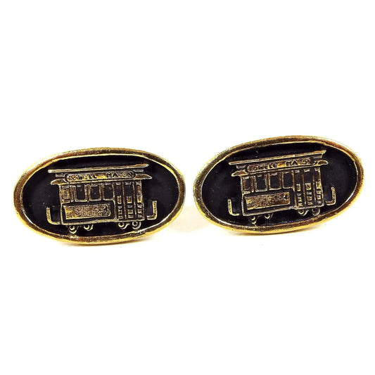 Front view of the Mid Century vintage Powell and Mason cable car cufflinks. The metal is gold tone in color and they are oval in shape. There is a cable car design with Powell Mason on the top and black painted background.