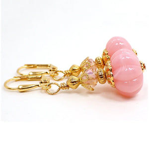 Side view of the handmade drop earrings with vintage lucite beads. The metal is gold plated in color. There is a new faceted glass crystal bead at the top in light pink. The bottom bead is pumpkin shaped and is light pink lucite with gentle swirls of white here and there.