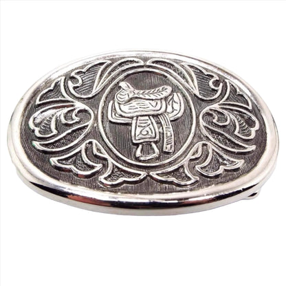 Front view of the retro vintage Avon belt buckle. The metal is silver tone in color with gray background. It's oval in shape with a horse saddle in the middle surrounded by a bell type floral design.