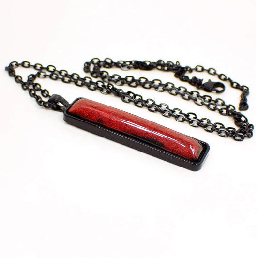 Angled side view of the handmade Goth resin pendant necklace. The chain and setting are black coated with a lobster claw clasp at the end. The pendant is a long bar shape with a rich shimmery red domed resin cab that has hints of black here and there around the edge.