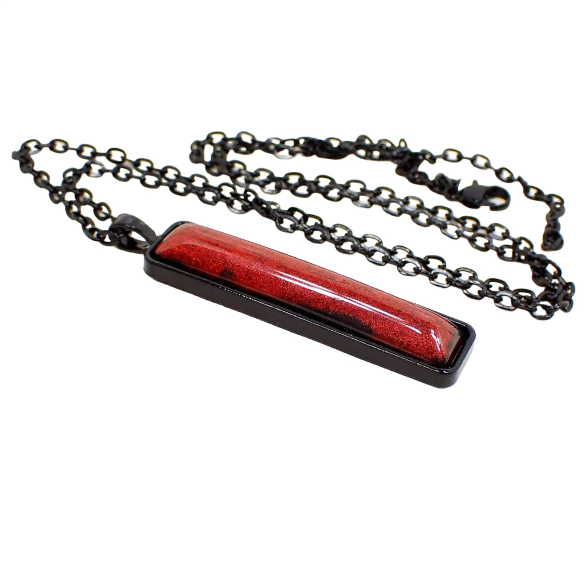 Angled side view of the handmade Goth resin pendant necklace. The chain and setting are black coated with a lobster claw clasp at the end. The pendant is a long bar shape with a rich shimmery red domed resin cab that has hints of black here and there around the edge.