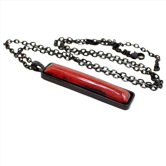 Angled side view of the handmade Goth resin pendant necklace. The chain and setting are black coated with a lobster claw clasp at the end. The pendant is a long bar shape with a rich shimmery red domed resin cab that has hints of black here and there around the edge.