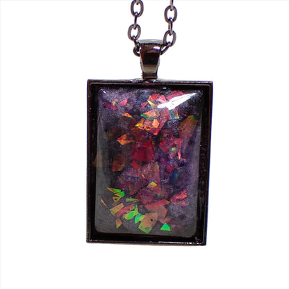 Enlarged front view of the handmade resin pendant. The metal is gunmetal gray in color. The resin is pearly dark purple with chunky iridescent glitter that flashes and shows different colors as it moves around in the light.