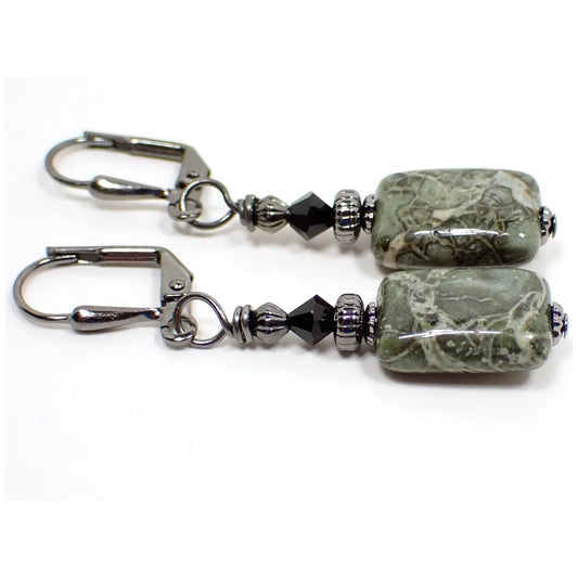 Photo of the handmade green rhyolite gemstone earrings. The metal is gunmetal gray in color. There are black faceted glass crystal beads at the top. The bottom gemstone beads are puffy square in shape and have a chunky pattern with shades of green, cream, and hints of brown.