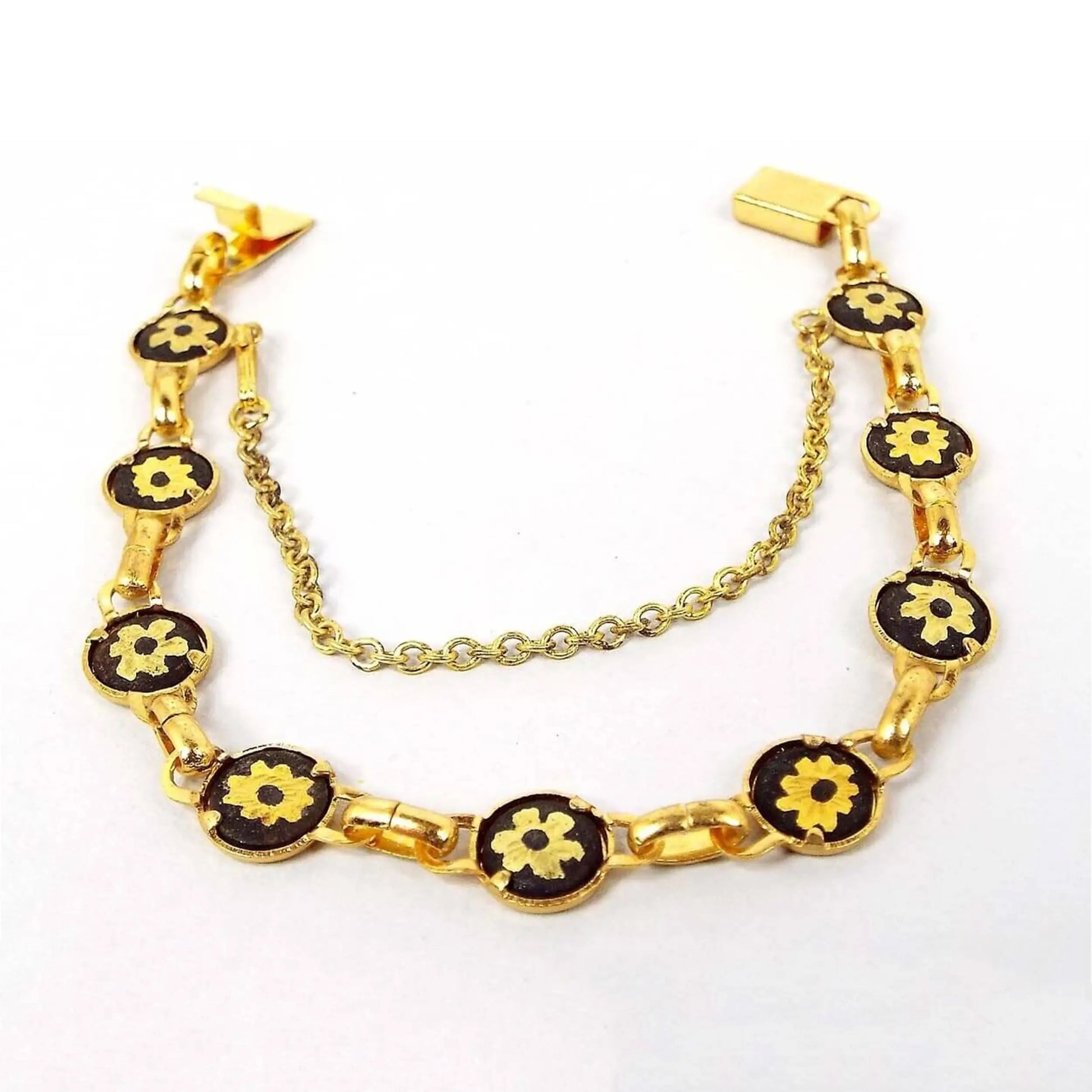 Front view of the retro vintage Damascene style bracelet. It is gold tone in color. The round links are painted black and have a gold tone flower design in the middle. There is a box clasp at the end and a safety chain with a hinged clip clasp as well.