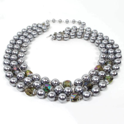 Front view of the multi strand Mid Century vintage gray faux pearl and crystal beaded necklace. There are three strands of beads. The strands mostly have gray imitation faux pearls with some ab brownish gray crystal beads towards the bottom of the necklace. There is a hook clasp at the end.