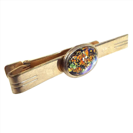 Front view of the Mid Century vintage tie clip with foil glass cab. The metal is gold tone in color. There is an oval glass cab in the middle with a metallic foil background that has shades of orange, green, yellow, and black.