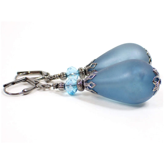 Side view of the large handmade teardrop earrings with vintage lucite beads. The metal is gunmetal gray in color. There are light blue faceted glass crystal beads at the top. The bottom lucite beads are wide teardrop shaped and have a matte frosted country blue color.