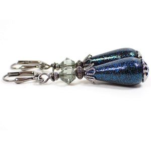 Side view of the handmade glitter teardrop earrings. The metal is gunmetal gray in color. There are light bluish gray faceted glass beads at the top. The bottom vintage acrylic beads are teardrop shaped, black in color, and have teal blue glitter on the outside.