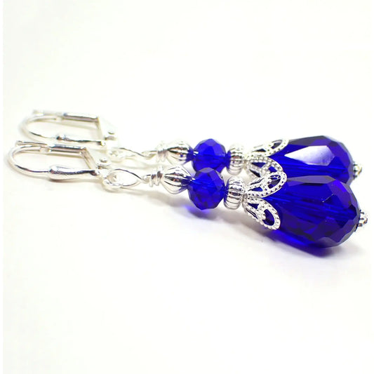 Angled view of the Victorian style handmade crystal glass teardrop earrings. The metal is silver plated in color. There is a faceted rondelle bead at the top and a faceted teardrop bead at the bottom. Both beads are a bright cobalt blue color.