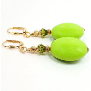 Angled view of the large handmade acrylic drop earrings. The metal is gold plated in color. There is a faceted green glass crystal bead at the top and a large oval vintage acrylic bead in lime green on the bottom.
