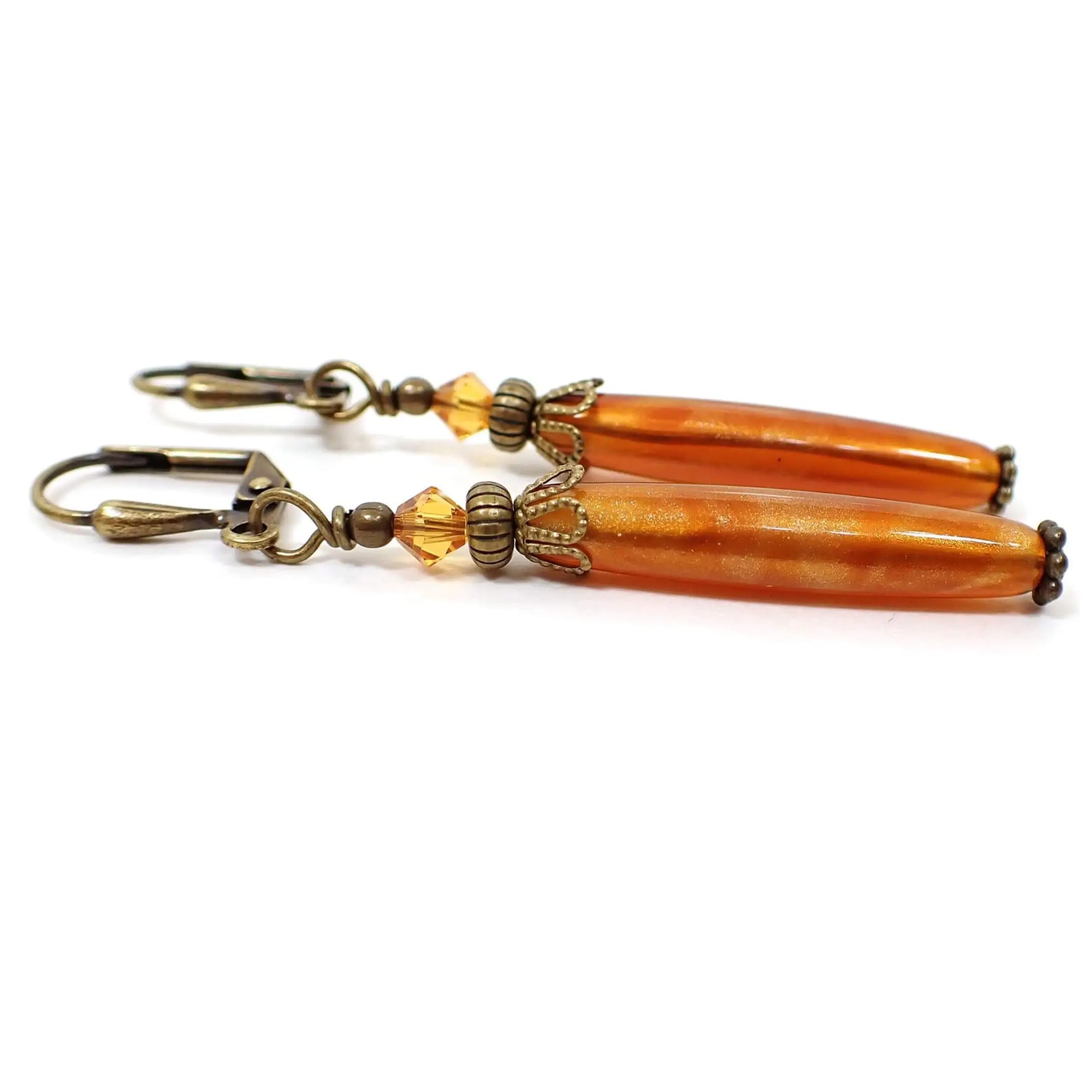 Side view of the handmade lucite stick earrings. The metal is antiqued brass in color. There are faceted glass crystals in a lighter orange at the top. The bottom lucite beads are round stick shaped and have orange marbled with metallic gold colors.