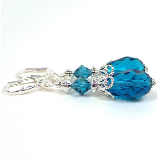 Side view of the handmade teardrop earrings. The metal is silver plated in color. There are teal blue faceted glass crystal bicone beads at the top and teardrop shaped beads at the bottom.