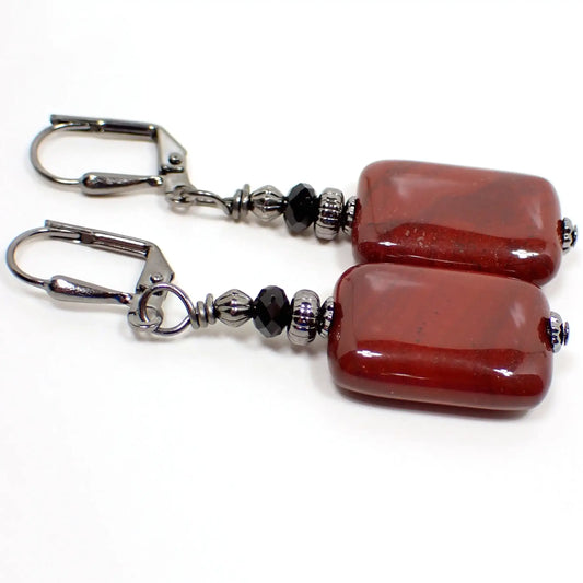 Angled view of the handmade red jasper earrings. The metal is gunmetal gray in color. There are black faceted glass crystal beads at the top. The bottom jasper bead is puffy rectangle shaped with rounded corners. It's primarily a dark brick red in color with a few areas of other colors marbled in.