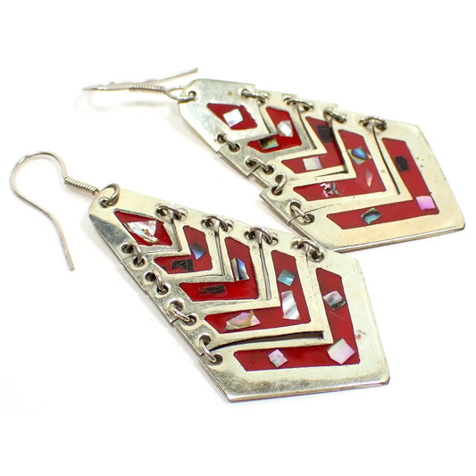 Enlarged angled view of the Southwestern retro vintage dangle earrings. The metal is silver tone plated in color. There are articulated pieces joined by a jump ring on each side with all the pieces together forming an angled teardrop style shape. Each piece is V or chevron shaped with red enamel in the middle and small pieces of inlaid abalone shell.