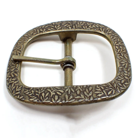 Top view of the retro vintage Burgos belt buckle. It has a oval like design with open middle showing a bar in the center. There is a leaf like pattern all the way around the buckle. It is antiqued brass in color with some darkened areas on the center bar.