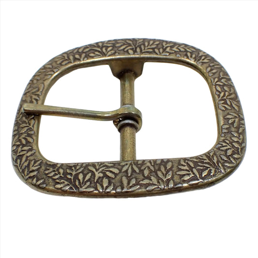 Top view of the retro vintage Burgos belt buckle. It has a oval like design with open middle showing a bar in the center. There is a leaf like pattern all the way around the buckle. It is antiqued brass in color with some darkened areas on the center bar.