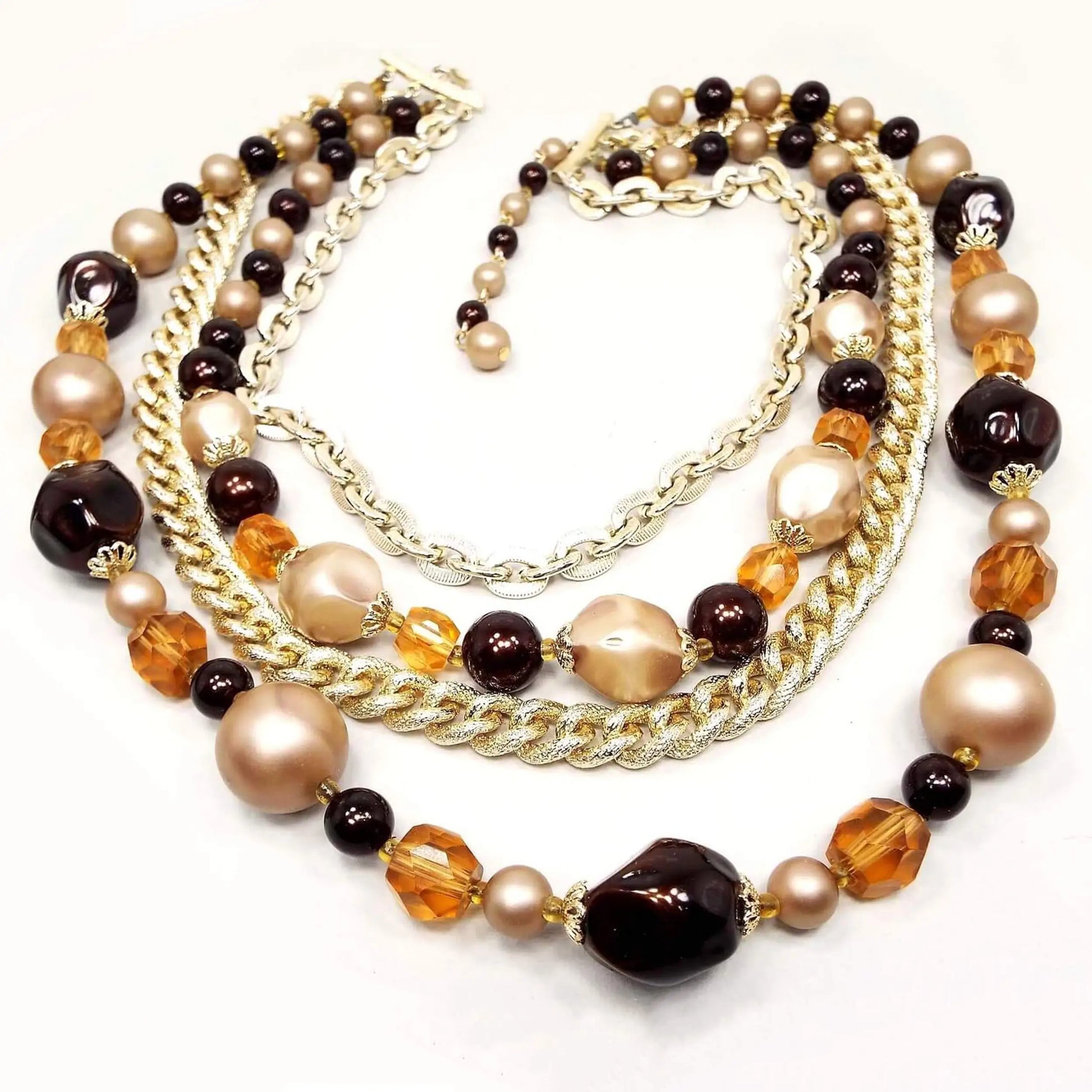 Front view of the Japanese Mid Century vintage multi strand beaded necklace. There are two strands of gold tone color chain and two strands of beads. The beads are chunky plastic, plastic faux pearl, and faceted glass beads in shades of orange, brown, and tan. There is a hook clasp at the end.