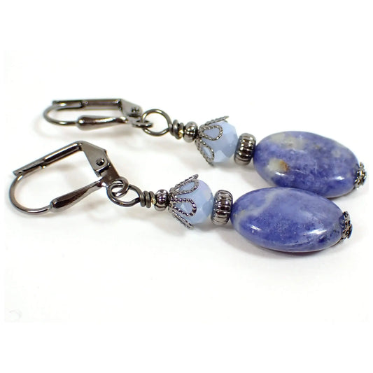Angled view of the handmade gemstone earrings. The metal is gunmetal gray in color. There are light blue faceted glass beads at the top. The bottom sodalite beads are puffy oval shaped and mostly denim blue in color with marbled areas of off white and a few specks of orange.