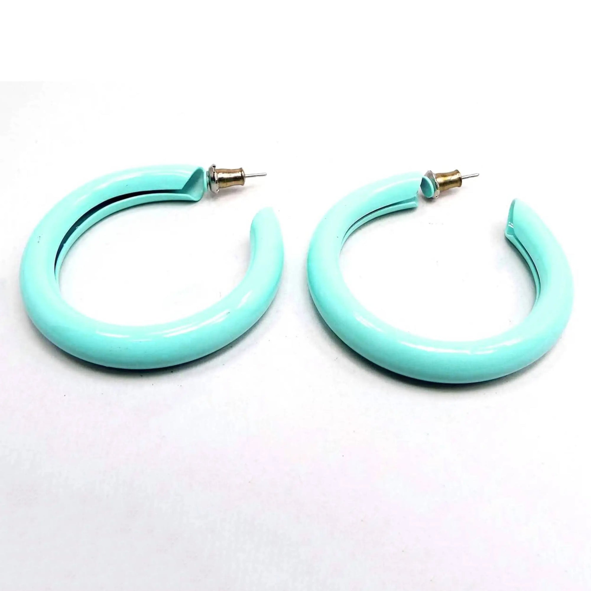 Angled side view of the retro vintage enameled hoop earrings. They are mostly sea foam blue green in color and have post ends with earring clutches attached.