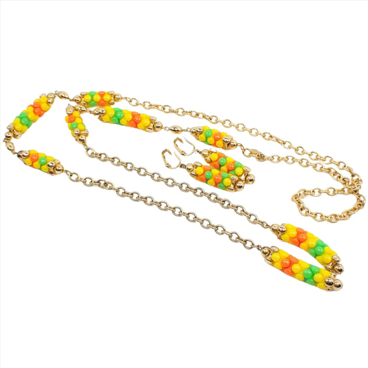 Angled view of the Mid Century vintage Sarah Coventry jewelry set. There is a lariat necklace with gold tone chain and plastic beads in several areas in yellow, orange, and green. There is also a matching pair of clip on earrings with beaded dangles in the same colors.