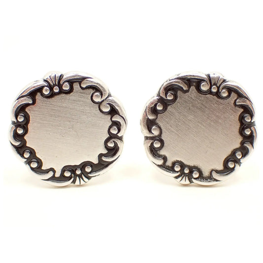 Front view of the Mid Century vintage Sarah Coventry cufflinks. They are round in shape and have a matte silver tone front with antiqued silver tone scroll edge.