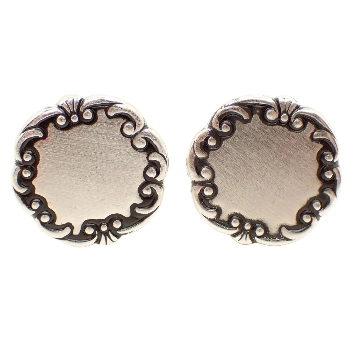 Front view of the Mid Century vintage Sarah Coventry cufflinks. They are round in shape and have a matte silver tone front with antiqued silver tone scroll edge.