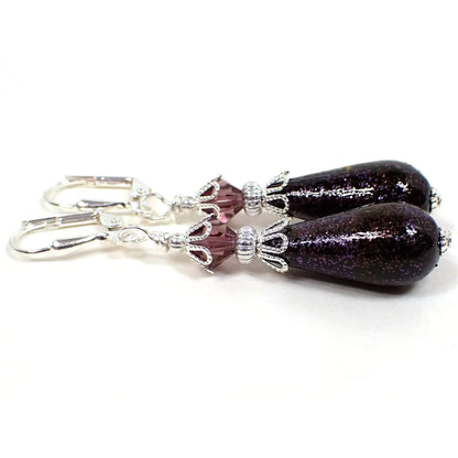 Side view of the handmade teardrop earrings with vintage German beads. The metal is silver plated in color. There are purple faceted glass crystal beads at the top. The bottom acrylic beads are teardrop shaped and are black with purple glitter on the outside.