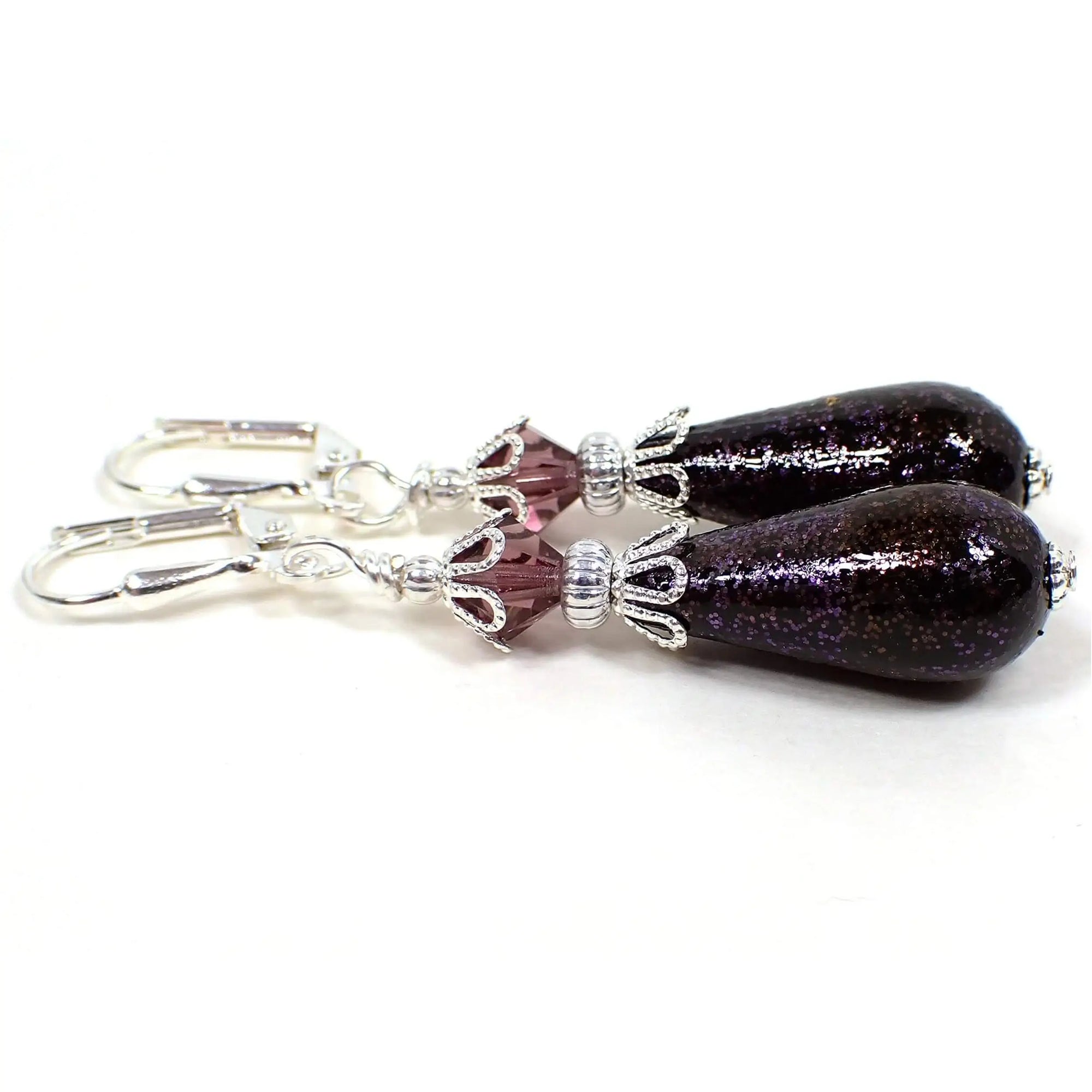 Side view of the handmade teardrop earrings with vintage German beads. The metal is silver plated in color. There are purple faceted glass crystal beads at the top. The bottom acrylic beads are teardrop shaped and are black with purple glitter on the outside.