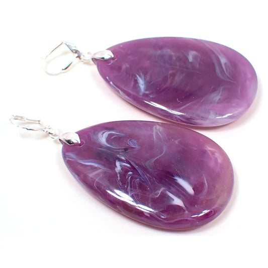 Angled view of the big heavy handmade teardrop earrings. The metal is silver plated in color. They are large teardrop shaped with shades of purple with swirls of white marbled in.