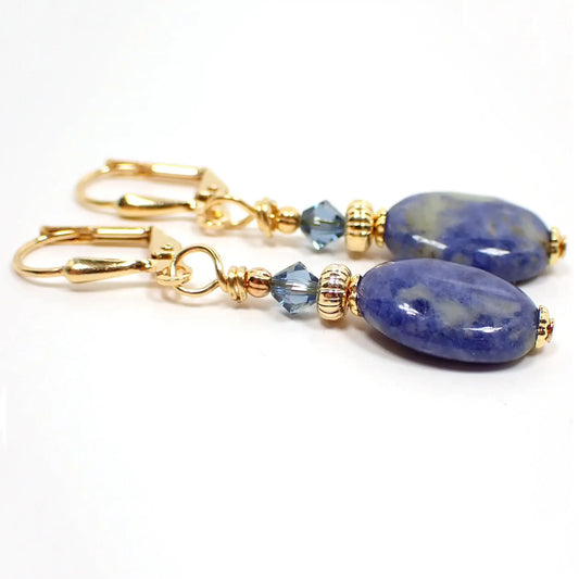 Photo of the handmade sodalite gemstone earrings. The metal is gold plated in color. There are small faceted glass crystal blue beads at the top. The bottom gemstone beads are puffy oval shaped and are denim blue with marbled areas of cream and brown colors.