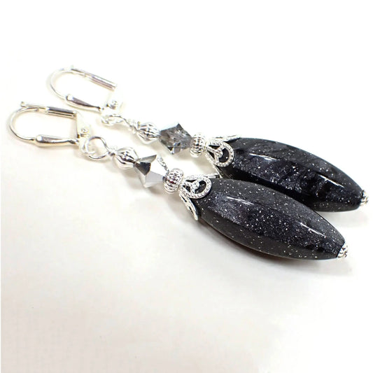 Angled view of the large handmade drop earrings with vintage lucite beads. The metal is silver plated in color. There are faceted clear and metallic silver glass crystal beads at the top. The bottom lucite beads are a long faceted oval shape and are pearly dark gray in color with tiny flecks of glitter embedded in them.