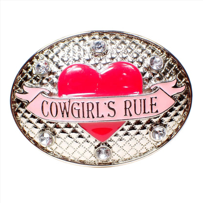 Front view of the pre owned Nocona belt buckle. It is silver tone plated in color and is an oval shape with a textured raised quilted design. There is a bright pink enameled heart in the middle with a light pink banner across it that says "Cowgirl's Rule." There are round clear rhinestones around the top and the bottom of the belt buckle.