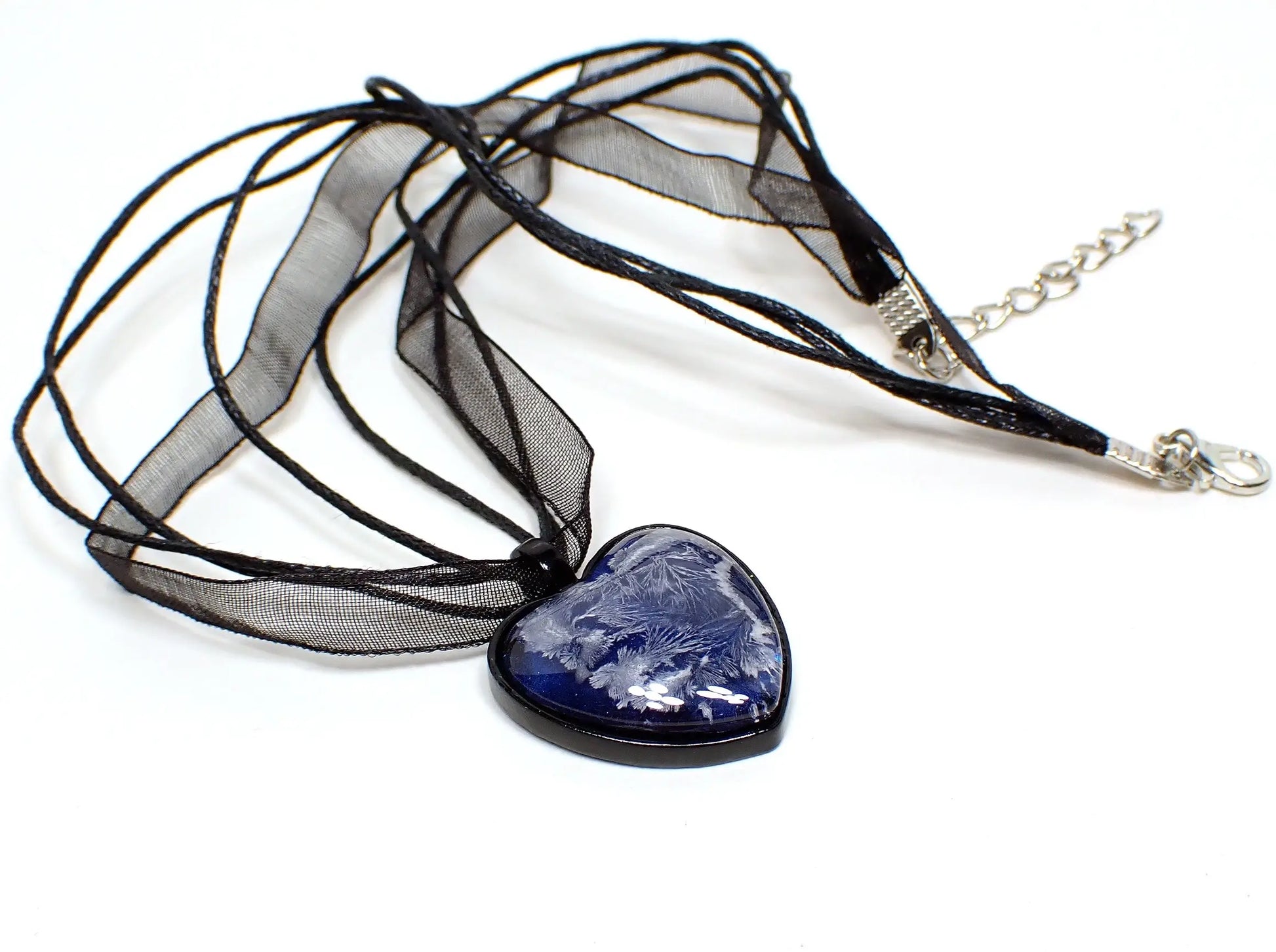Top view of the handmade cold hearted black heart pendant necklace. The necklace is black with three strands of faux leather cord and a strand of organza ribbon. There is a lobster claw clasp and extender chain at the end. The heart pendant is black coated with iridescent blue resin and a white abstract frost like design through the resin.