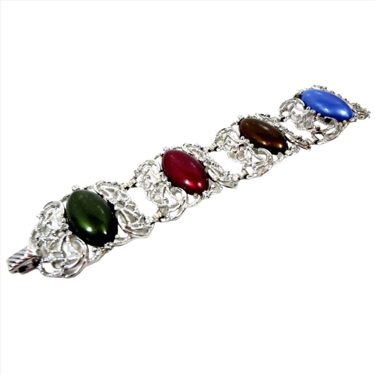 Top view of the Mid Century vintage Sarah Coventry link bracelet. It is silver tone in color with wide curved filigree links. Each link has a large oval lucite cab. Each one is a different color. There is green, red, brown, and blue. There is a snap lock clasp at the end.