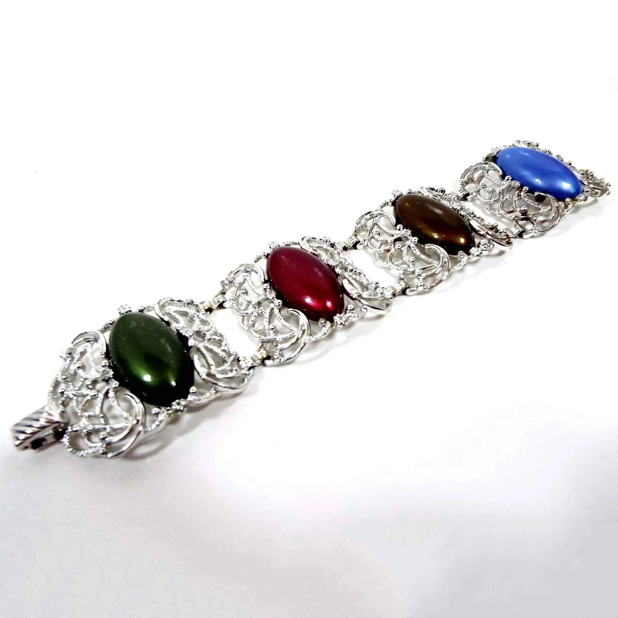 Top view of the Mid Century vintage Sarah Coventry link bracelet. It is silver tone in color with wide curved filigree links. Each link has a large oval lucite cab. Each one is a different color. There is green, red, brown, and blue. There is a snap lock clasp at the end.