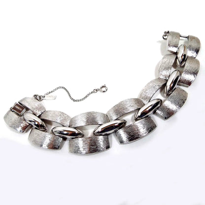 Top view of the Mid Century vintage Monet link bracelet. It is silver tone in color with wide brushed matte links with shiny parts in the middle. The main part of the links are open curved square shape with long ovals in the middle. There is a snap lock clasp and a small safety chain with spring ring clasp. \