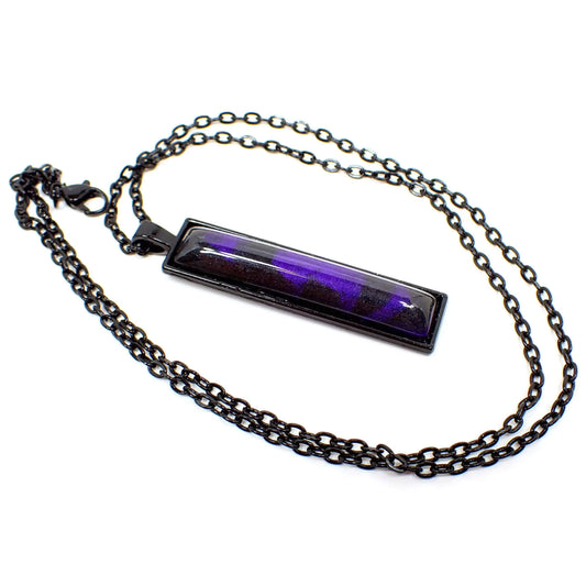 Angled front view of the Goth Handmade Black and Purple Resin Bar Pendant. The chain and setting are black coated metal. The pendant has a long rectangle bar shape and a domed resin cab with marbled areas of pearly black and purple colors.