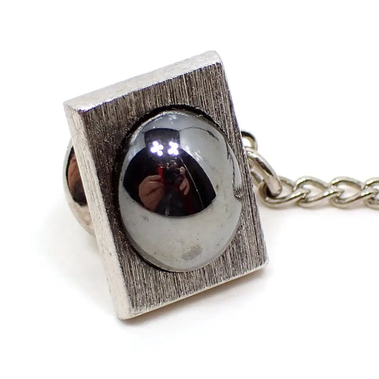 Enlarged angled front view of the Sarah Coventry tie tack. It has a rectangle shape with a brushed matte silver tone appearance. There is a domed oval hematite cab in the middle that is a dark metallic gray. The hematite is shiny and you can see my reflection taking the photo in it.