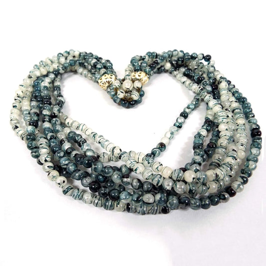 Front view of the Mid Century vintage Hong Kong multi strand necklace. It has seven strands of smaller sized round white beads that have a swirl design of dark green with hints of blue for an overall dark teal green appearance. There is a round beaded box clasp at the end.
