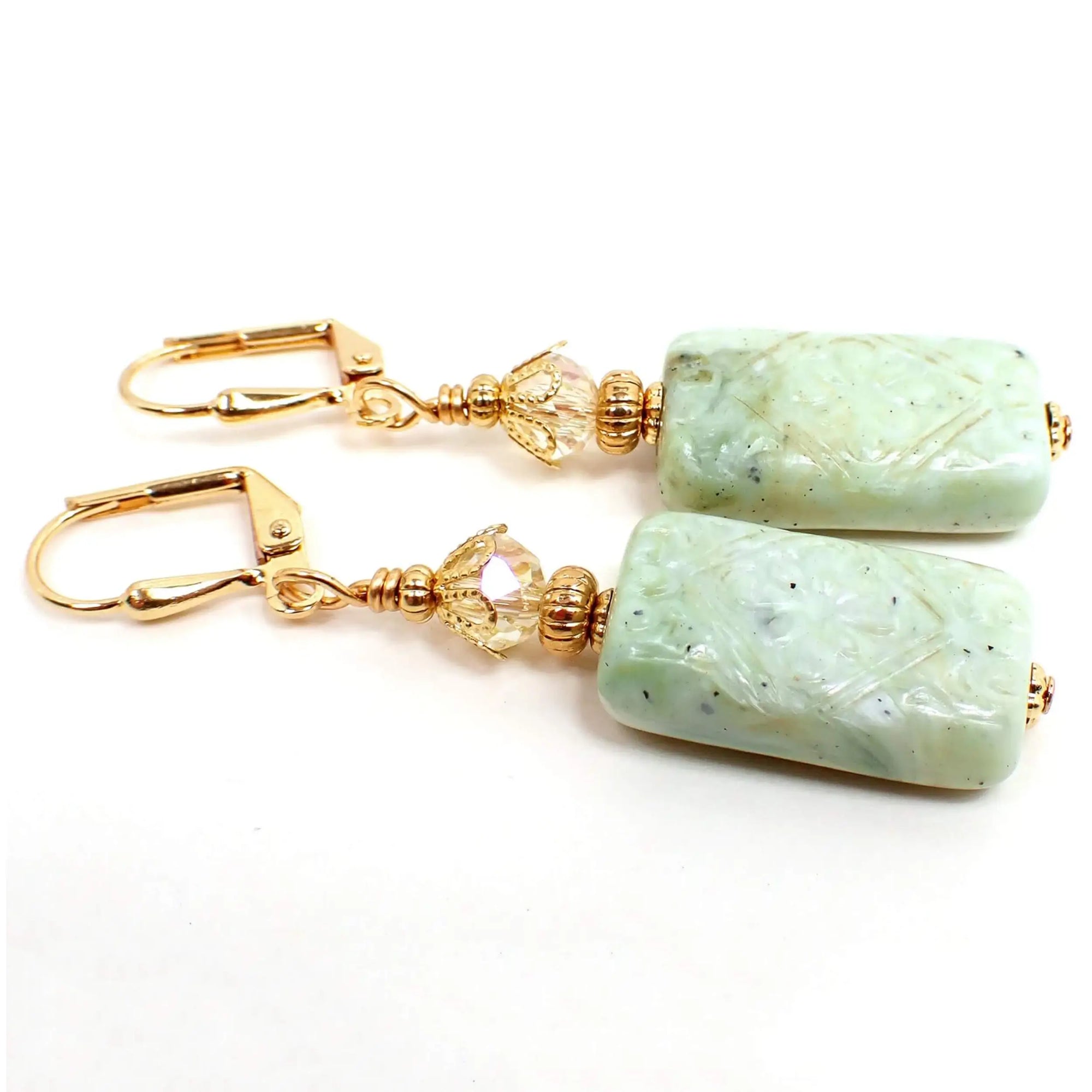 Angled view of the handmade rectangle drop earrings. The metal is gold plated in color. There are light yellow faceted glass crystal beads at the top. The bottom rectangle beads are acrylic with a textured floral pattern and mottled shades of light green.