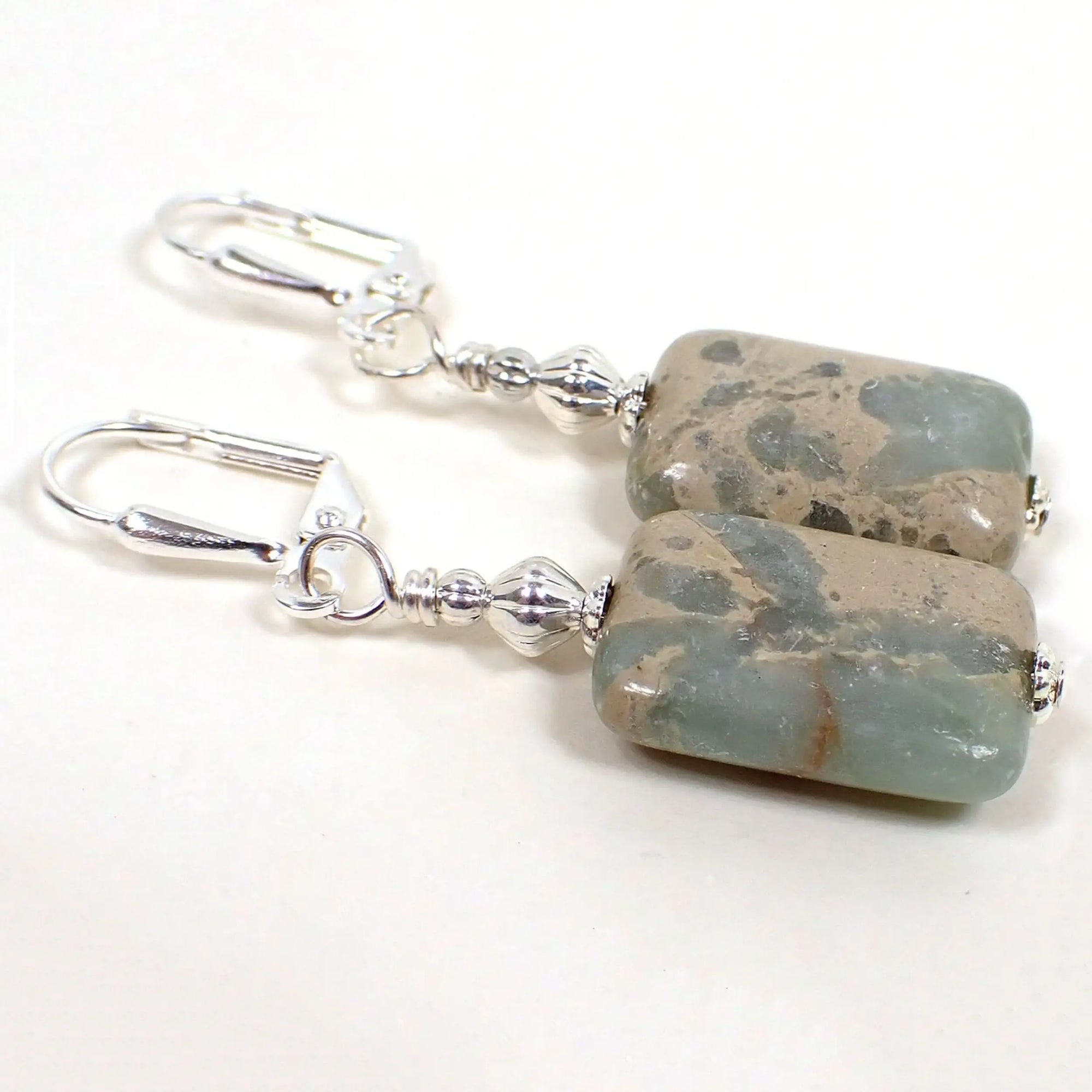 Angled view of the handmade gemstone earrings. The metal is silver plated in color. There are impression jasper beads at the bottom in a puffy rectangle shape. Impression jasper is manmade stone and has shades of light aqua blue marbled with shades of tan, cream, and may have a reddish line here or there.