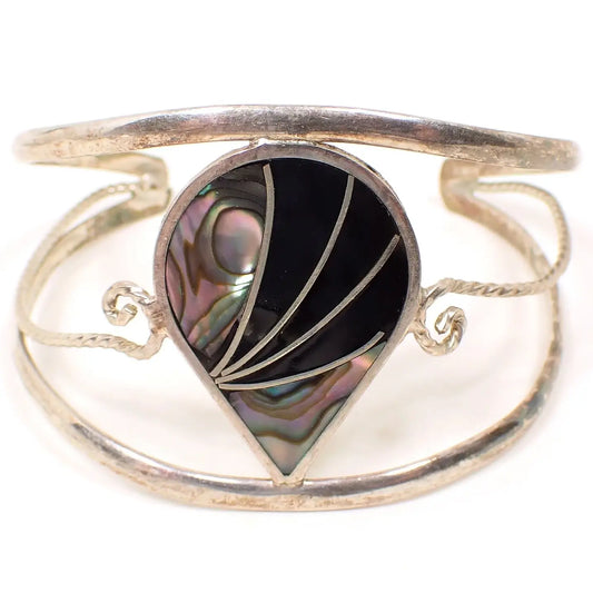 Enlarged front view of the Alpaca Mexican bracelet. The metal is silver tone in color. There are three rounded bands with open spaces between them making up the cuff part of the bracelet. The top and bottom are curved. The middle one has a twisted design with curls at the end. In the middle is a teardrop shape with inlaid abalone shell and black enamel with silver tone color metal curves between the areas.